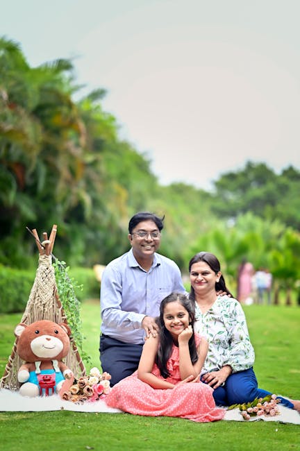 A family of four outdoors on a grassy area during daytime, with a lush green background of trees and foliage. The father, wearing glasses and a light blue shirt, is seated behind his wife and daughter, with his arm around their shoulders, smiling at the camera. The mother, dressed in a patterned blouse, sits next to her daughter, who appears to be around six years old and is wearing a pink dress with small white polka dots, smiling happily. The daughter is sitting on the grass, leaning slightly against her mother. To the left, there is a small decorative setup featuring a plush teddy bear with a friendly face, a miniature teepee tent made of natural-colored fabric with wooden sticks, and some floral embellishments, all arranged on the grass. The scene has a bright, natural light atmosphere that highlights the vibrant colors of the family’s clothing and the greenery surrounding them, creating a warm and cheerful mood suitable for a family-friendly context related to waste management or outdoor community activities.