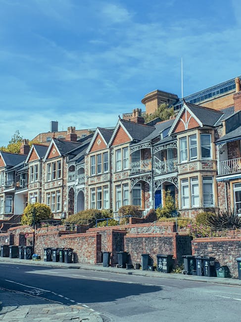 A row of Victorian-style terraced houses with decorative white wrought iron balconies and bay windows, set along a residential street with a brick retaining wall and a series of black waste bins lined up along the pavement. The houses have red brick facades with contrasting white trim and gabled roofs, and the scene includes greenery such as small bushes and trees near the front of the properties. In the background, taller modern buildings can be seen under a bright blue sky with scattered clouds. The image captures a typical urban residential area where private rubbish collection or on-site waste clearance services, like those offered by Rubbish Clearance Haringey, might be employed to manage household waste, especially given the visible presence of waste bins intended for refuse collection.