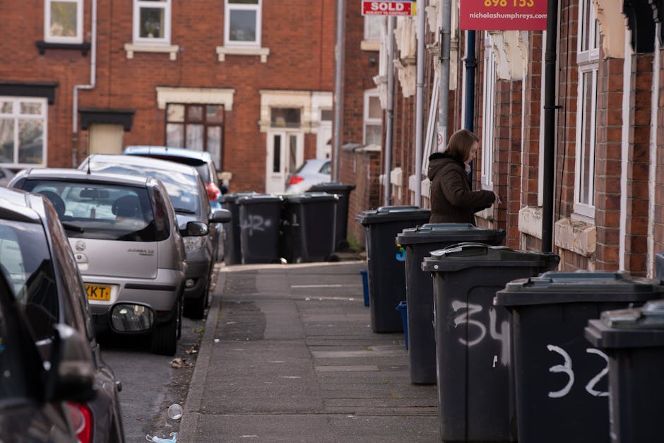 The image depicts a residential street scene with multiple dark grey wheelie bins lined up along the pavement in front of brick terraced houses, some with graffiti markings on their sides. A woman dressed in a brown coat is standing next to one of the bins, appearing to be opening or closing the lid. The pavement is slightly worn with visible grime and a few scattered pieces of litter. Several parked cars, including a silver hatchback in the foreground, occupy the street alongside the bins, with more vehicles visible further down the road. The background features brick houses with white window frames and some signs, including one advertising a property sale, attached to the buildings. The scene is lit by natural daylight, with an overcast sky casting diffuse light across the area, reflecting typical urban waste management practices where residents or private rubbish removal services, such as Rubbish Clearance Haringey, handle waste collection outside individual property boundaries in an organized manner.